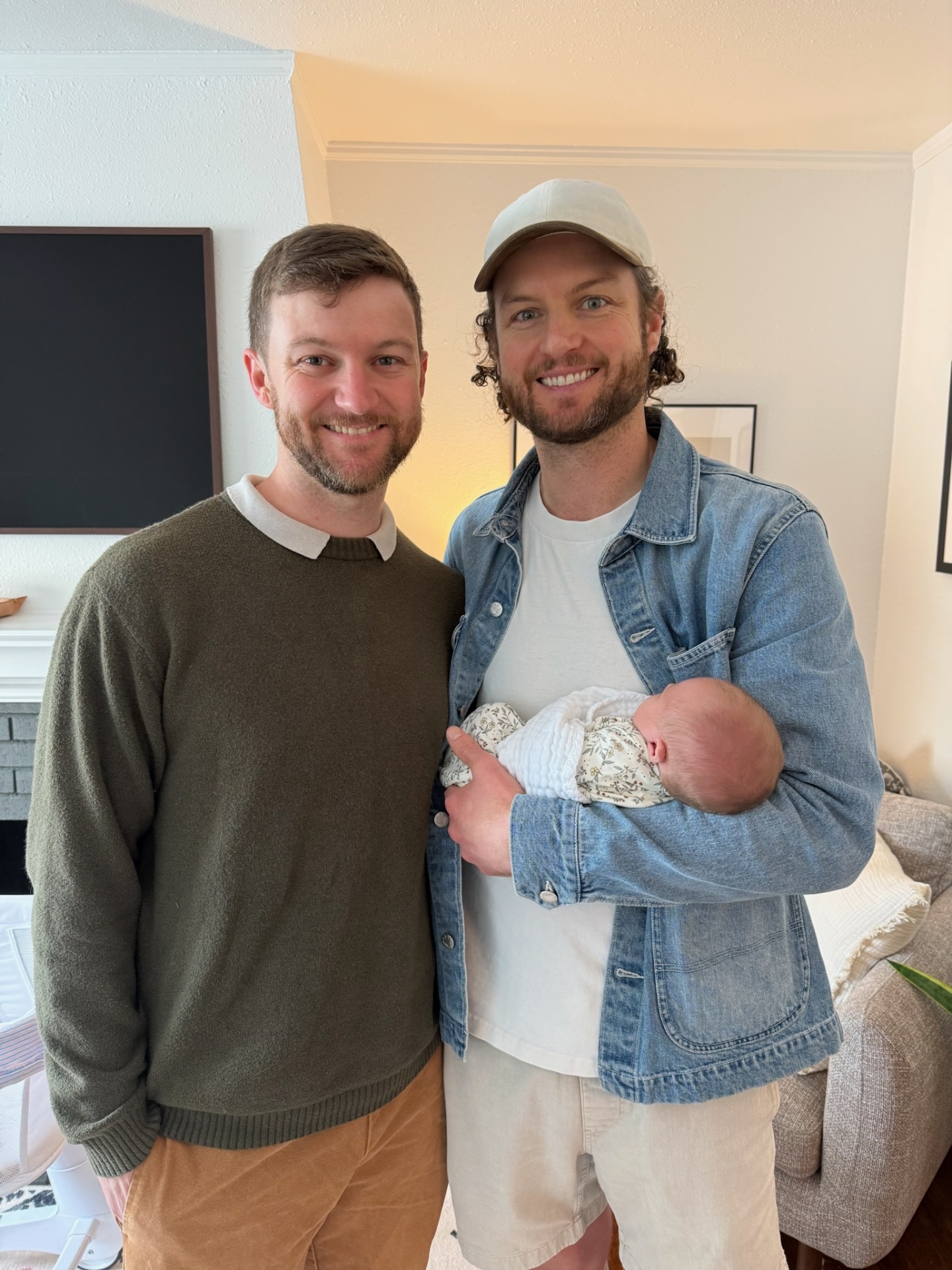 Two brothers stand together, one cradling a newborn baby in his arms.