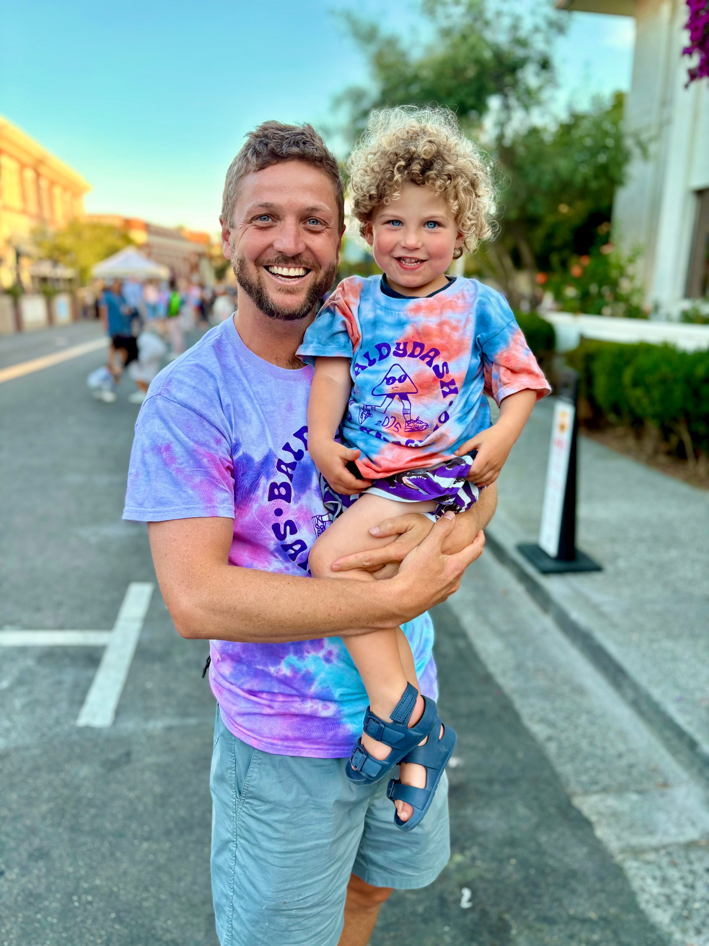 A father holds his young son on a summer evening — both grinning.